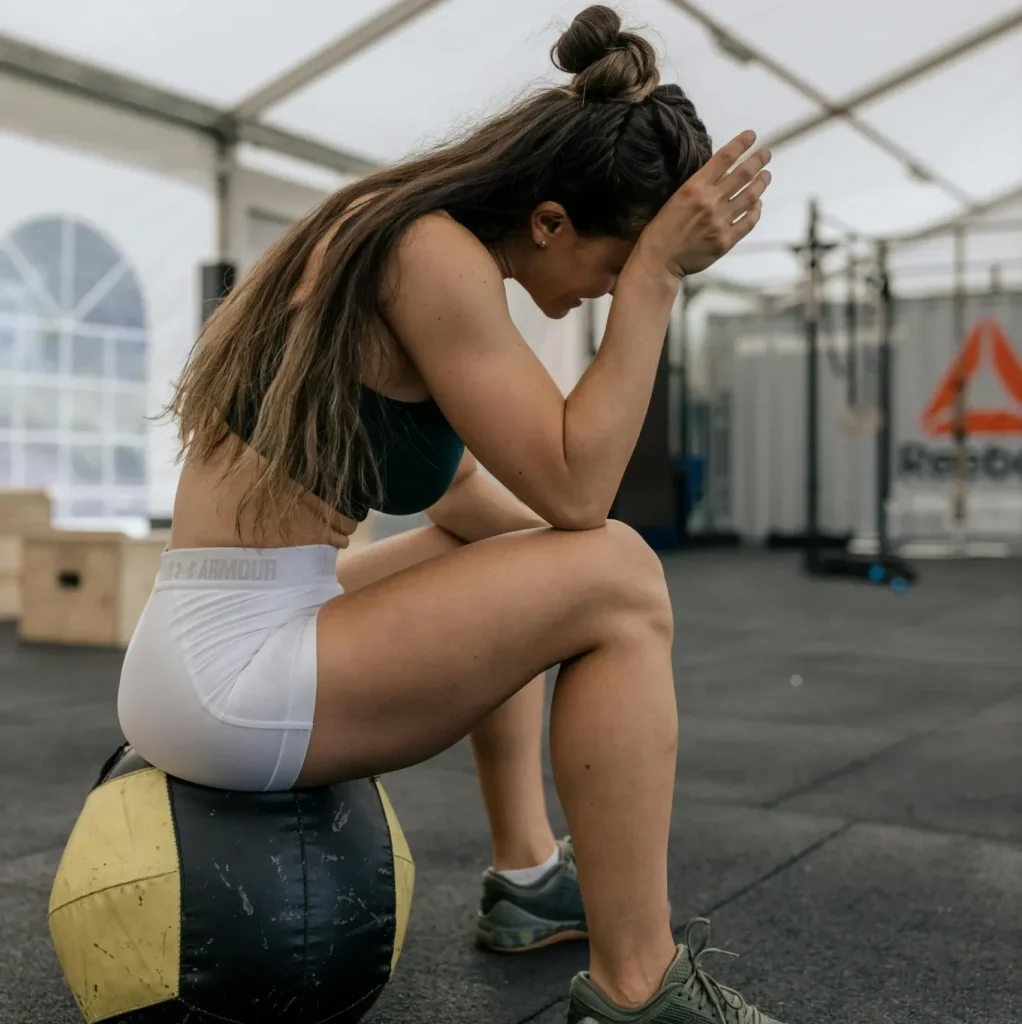 Athlète féminine en récupération après un entraînement intensif de crossfit, assise sur un medicine ball dans une salle de sport couverte. CBD prenium d'ardèche.
